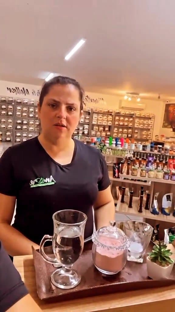 Atendente da Amore no balcão de madeira, com copos de chá e detox na bandeja e as prateleiras ao fundo
